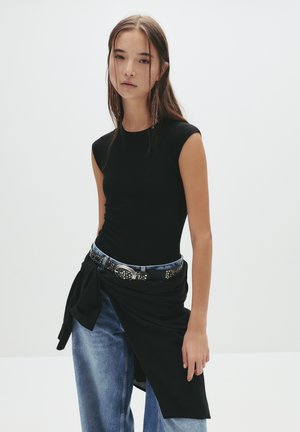 Svart åtsittande ärmlös topp i kombination med högmidjade denimjeans. Outfiten har ett knutet svart tygaccent och ett nitarbälte.