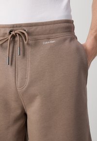 Brązowe dresy wykonane z miękkiego materiału, z elastycznym paskiem z możliwością regulacji, bocznymi kieszeniami oraz logo Calvin Klein z przodu.