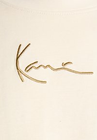 Kremo spalvos marškiniai, dekoruoti auksiniu siuvinėtu parašu, pagaminti iš lygaus, lengvo audinio ir turintys prigludusį, apvalų kaklo iškirpimą.