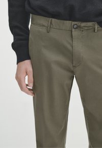 Pantaloni verde oliva con una trama liscia, dotati di chiusura con bottone e tasche frontali, abbinati a un maglione scuro.