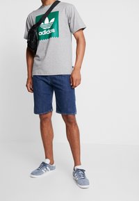 Man i grå Adidas t-shirt, denimshorts, ljusblå sneakers och svart axelremsväska mot en vanlig vit bakgrund.