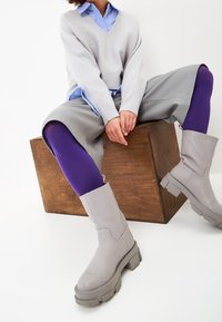 Suéter gris sobre una camisa azul claro, pantalones cortos sintéticos grises, mallas moradas y botas gruesas grises con suela texturizada, sentado en un bloque de madera.