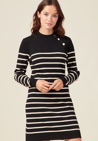 Abito a maglia a strisce bianche e nere con colletto alto e polsini a coste. Presenta tre bottoni sulla spalla sinistra. Texture in maglia morbida.