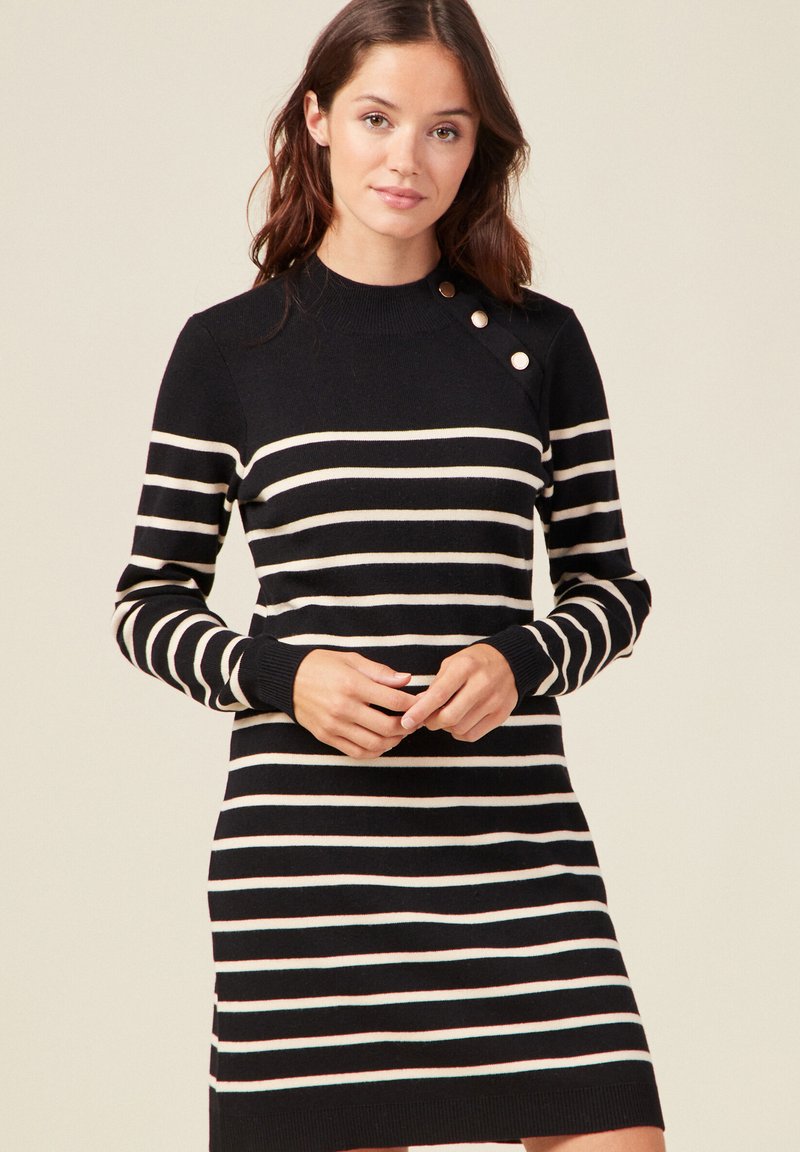 Abito a maglia a strisce bianche e nere con colletto alto e polsini a coste. Presenta tre bottoni sulla spalla sinistra. Texture in maglia morbida.