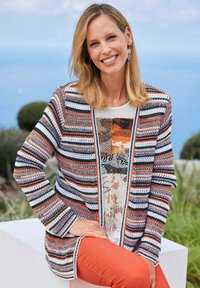 Gebreide cardigan met horizontale strepen in oranje, marineblauw en wit. Gelaagd over een crèmekleurige top met abstracte patronen. Effen oranje broek.