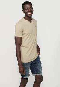 Beige színű, rövid ujjas henley póló finom logóhímzéssel, kék farmer cargo rövidnadrággal párosítva, amelynek felhajtott szárai és többzsebes kialakítása van.