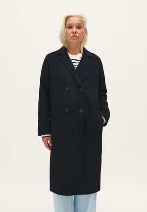 Cappotto navy doppiopetto in misto lana, con grandi bottoni, colletto ampio e tasche laterali. Indossato sopra una camicia a righe e jeans chiari.