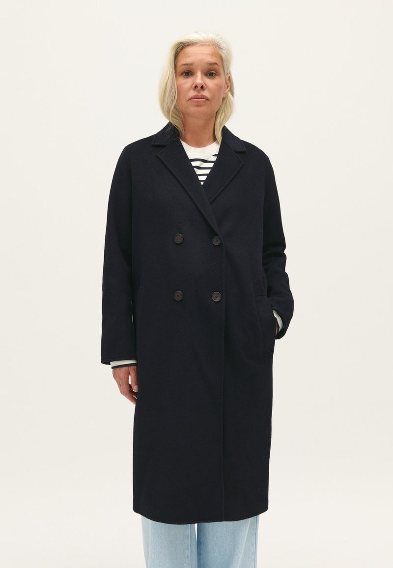 Cappotto navy doppiopetto in misto lana, con grandi bottoni, colletto ampio e tasche laterali. Indossato sopra una camicia a righe e jeans chiari.