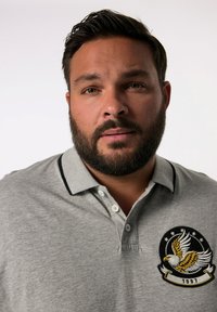 Grå polotröja med svart krage, prydd med en broderad örnlogotyp och texten "1997" på vänster bröstområde. Slät stickad textur.