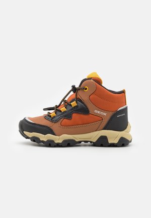Barns brun og orange vandrestøvle med sort tåbeskytter, beige robust sål og elastiske snøretog, mærket "Geox Amphibiox" på siden.