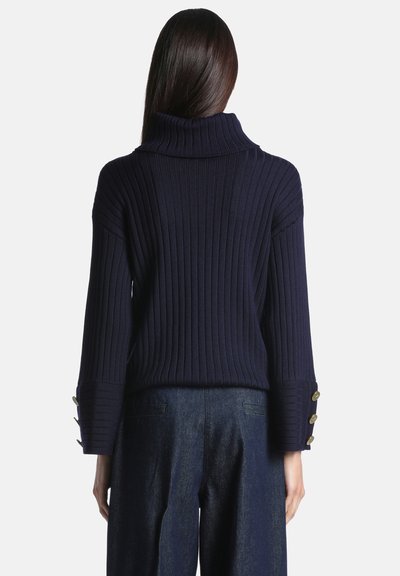 Femme portant un pull à col roulé côtelé bleu foncé avec des poignets boutonnés et un pantalon large sombre, vue de dos.