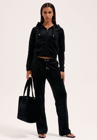 Kvinna klädd i svart velourhuva och byxor, stora hoopsörhängen, håller en svart tygpåse och har på sig svarta flip-flopsandaler, står.