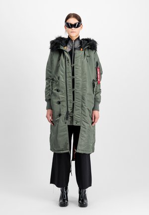 Olive green padded long coat with a fur-trimmed hood, button closures, and a side zip pocket. Worn over black wide-leg pants and black boots.