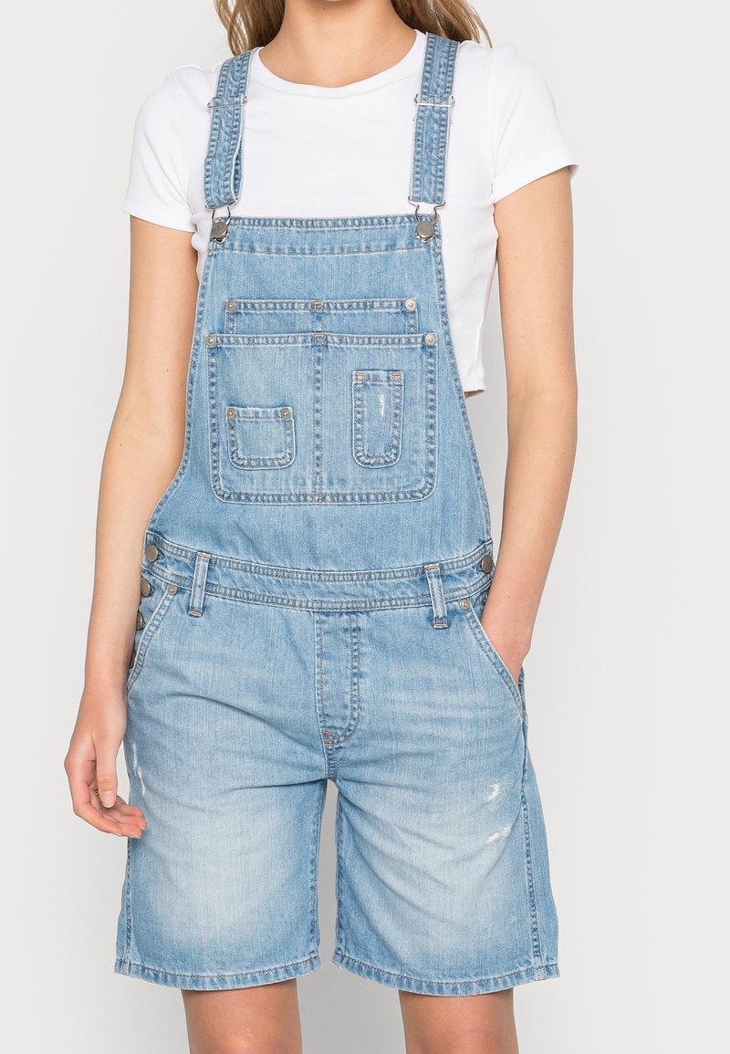 Salopette en denim bleu clair avec une coupe décontractée, bretelles réglables et plusieurs poches avant. Longueur short avec détails usés.