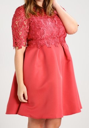 Mujer con un vestido coral hasta la rodilla con mangas cortas de encaje y detalles florales de encaje en el corpiño.