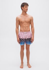 Λεπτός άνδρας όρθιος, ξυπόλητος, φορώντας ροζ και μπλε μαγιό με κορδόνι και την επιγραφή "SQUARED", κοιτάζει προς τα αριστερά του σε απλό λευκό φόντο.