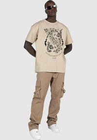 Beige grafisk t-skjorte med svart blomsterdesign, kombinert med brune cargo-bukser med sidelommer og hvite sneakers. Modell med solbriller.