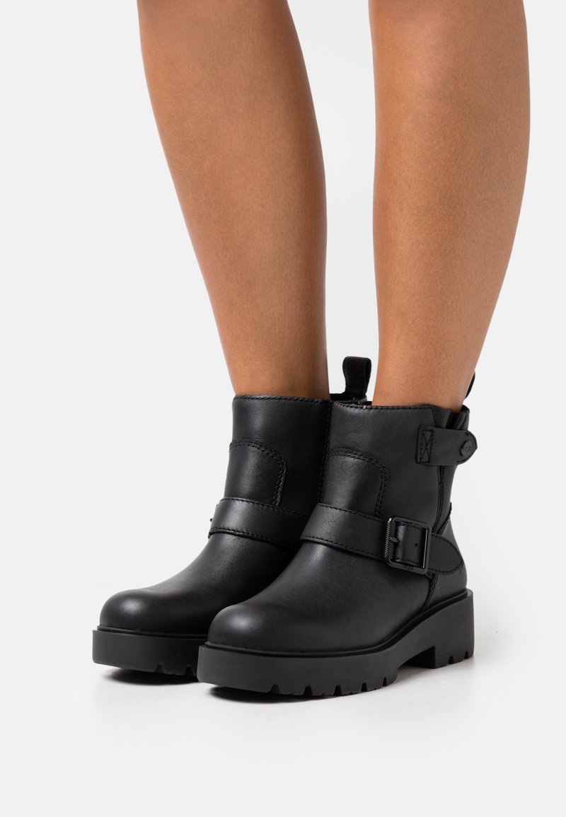 Jambes portant des bottines en cuir noir avec une large lanière, un détail de boucle et des semelles épaisses sur un fond clair.