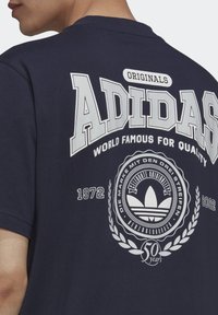 Mörkblå t-shirt med en stor vit Adidas-logotyp och texten "Originals" på baksidan, med en cirkulär badge och årtal.