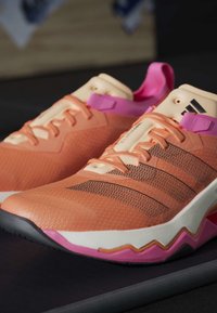 Baskets Adidas orange et rose avec des bandes noires, des lacets couleur pêche et des semelles rembourrées reposant sur une surface sombre.