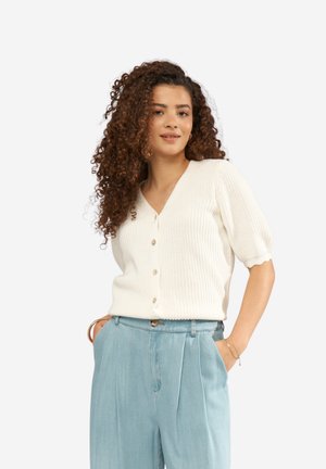 Młoda kobieta o kręconych włosach, ubrana w biały, zapinany na guziki sweter z krótkim rękawem i jasnoniebieskie spodnie z wysokim stanem, pozująca z rękami w kieszeniach.