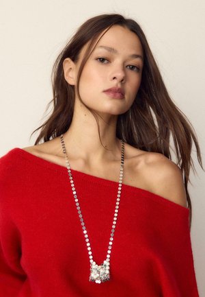 Giovane donna con lunghi capelli castani che indossa un maglione rosso scivolato sulle spalle e una lunga collana di paillettes argento su uno sfondo semplice.