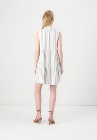 Robe blanche sans manches avec des rayures verticales bleues, un design en volants superposés et un col. Associée à des talons strappy marron clair.