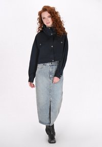 Korte zwarte jas met twee borstzakken en drukknoopsluitingen, gecombineerd met een lichtblauwe denimrok met een split aan de voorkant en zwarte laarzen.