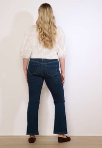 Žena s dlhými vlnitými blond vlasmi stojí chrbtom ku kamere, oblečená v bielej blúzke s nafúknutými rukávmi, tmavomodrých džínsoch a hnedých nazúvacích topánkach.