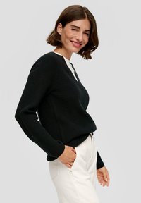 Schwarzer, gerippter Pullover mit V-Ausschnitt, langen Ärmeln, strukturiertem Stoff, kombiniert mit hellen Hosen. Minimalistisches Design, neutrale Farben.