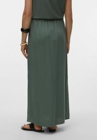 Robe maxi en tissu vert doux, dotée d'une taille élastique et d'un design fluide. Les accessoires comprennent un bracelet noir et des sandales.