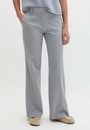 Kobieta ubrana w szerokie spodnie w jasnoszarym kolorze, beżowe lordsy i jasnoniebieski sweter dzianinowy z rękami w kieszeniach.