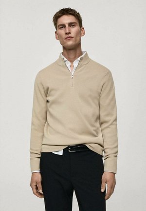 Beżowy sweter z zamkiem przy szyi, wykonany z miękkiego materiału, z długimi rękawami i luźnym krojem, noszony na białej koszuli z kołnierzykiem oraz czarnych spodniach.