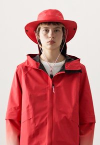 Veste imperméable rouge à manches courtes et capuche ajustable. Elle possède une fermeture éclair à l'avant et un cordon de serrage ; portée avec un chapeau à large bord assorti rouge.