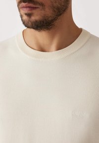 Pull en maille beige clair avec un col rond, présentant un motif texturé et un logo de marque subtil tissé dans le tissu.