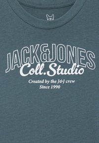Teal bomull T-skjorte med stor hvit tekst, "JACK & JONES Coll. Studio," og et lite grafisk logo nær halsen. Glatt tekstur.