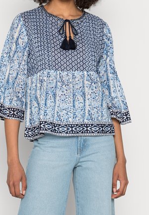 Femme portant un chemisier à manches longues bleu et blanc à motifs avec un col à nouer à pompons, associé à un jean bleu clair.