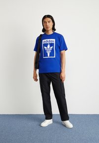 Blue cotton t-shirt with white graphic design featuring a torch and "adidas" logo. Paired with black cargo pants and white sneakers.
