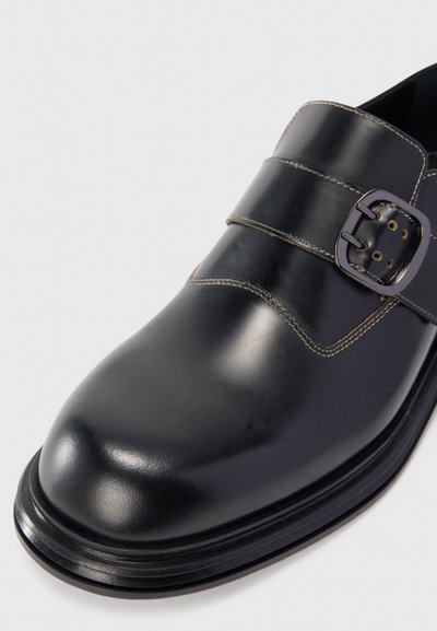Chaussure monk en cuir noir avec boucle en argent, bout arrondi et couture visible sur fond blanc.