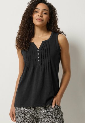 Vrouw met krullend haar, gekleed in een mouwloos zwart blouse met knopen en plooien, en zwart-witte patroonbroek, staand tegen een effen muur.