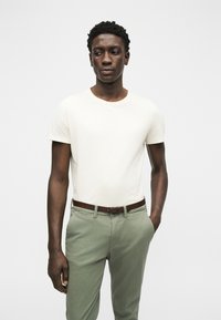 Άνδρας που φοράει ένα απλό λευκό κοντομάνικο μπλουζάκι, ανοιχτό πράσινο παντελόνι με καφέ ζώνη, στέκεται μπροστά σε απλό λευκό φόντο.