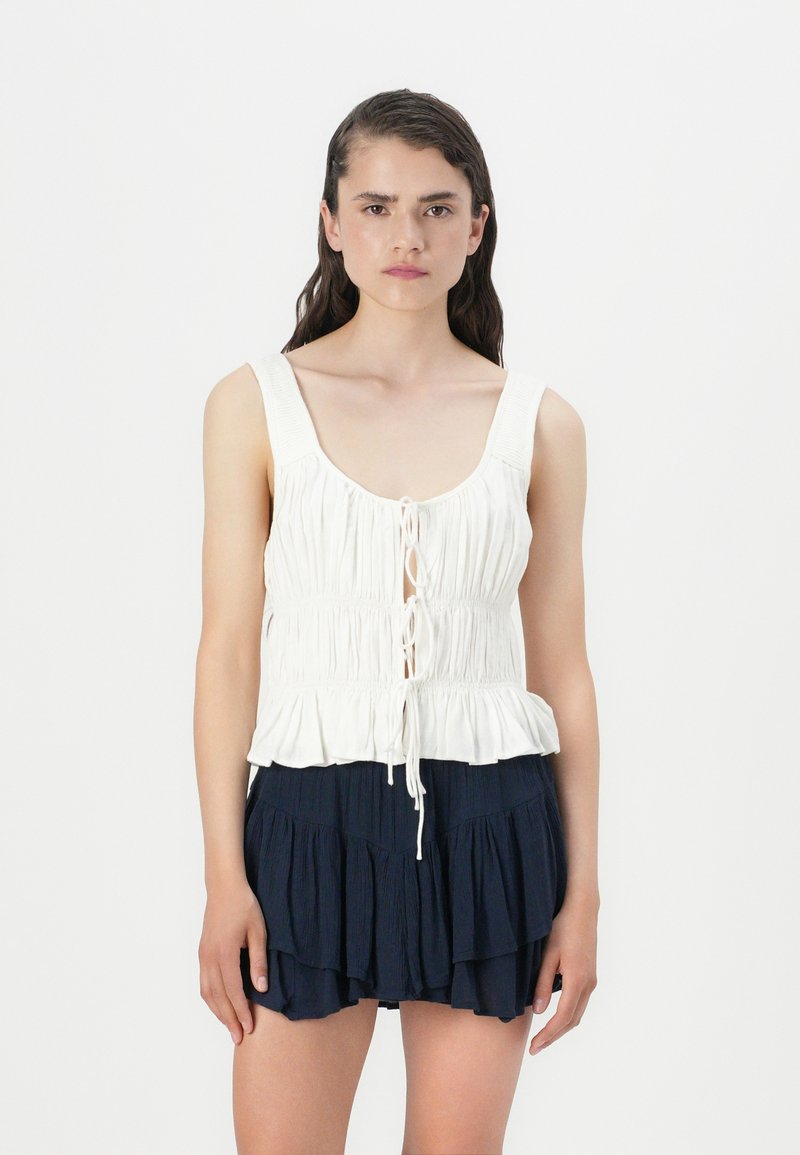 Top sans manches blanc avec une texture froncée et un devant à cordon de serrage, associé à un short bleu marine avec un design à volants en niveaux.