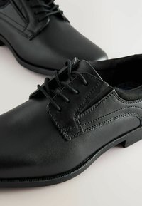 Chaussures de ville en cuir noir avec une finition lisse, présentant un design à lacets, des accents texturés et un talon bas. Détails de couture discrets visibles.