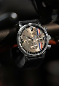 Zegarek Timex Marlin z srebrną stalową kopertą, złotą tarczą, czarnym skórzanym paskiem oraz czerwonymi, białymi i niebieskimi akcentami. Zegarek kwarcowy z chronografem.
