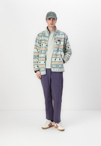 Veste en polaire avec des motifs géométriques en bleu, pêche et crème ; associée à un tee-shirt vert clair, un pantalon violet et des baskets blanches avec des rayures rouges.