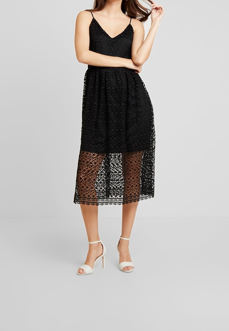 Svart spetsklänning med V-ringning, figurnära liv och midilängd A-linje kjol med ett genomskinligt lager. Kombinerad med vita högklackade skor.