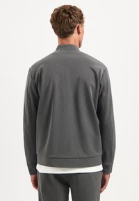 Grijze bomberjack met een ribbelende tailleband en manchetten, met een gladde textuur en minimalistisch ontwerp. Gemaakt over een wit shirt.