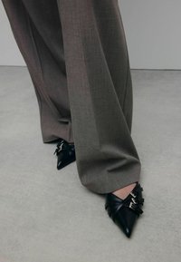 Chaussures noires à bout pointu en cuir texturé, avec des sangles et des boucles décoratives. Portées avec un pantalon gris ample et ajusté.