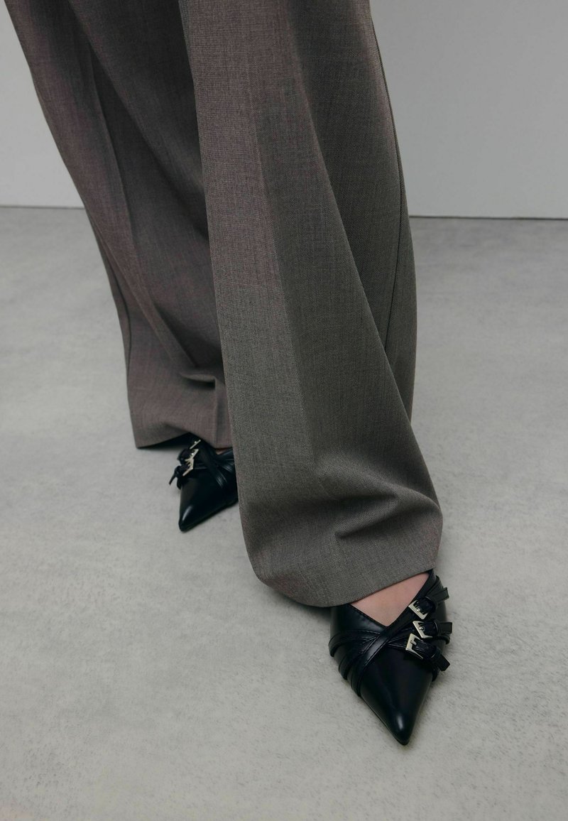 Chaussures noires à bout pointu en cuir texturé, avec des sangles et des boucles décoratives. Portées avec un pantalon gris ample et ajusté.