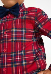 Rød ternet skjorte med blå, gul og hvide accenter, der har en laplomme og knaplukning. Sammen med en marineblå butterfly.